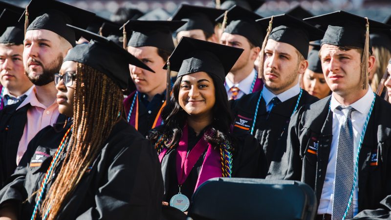 Two students sit among their graduating peers at the 2019 Undergraduate Commencement Ceremony, clapping and smiling. Photo by Erin Williams for Virginia Tech.
