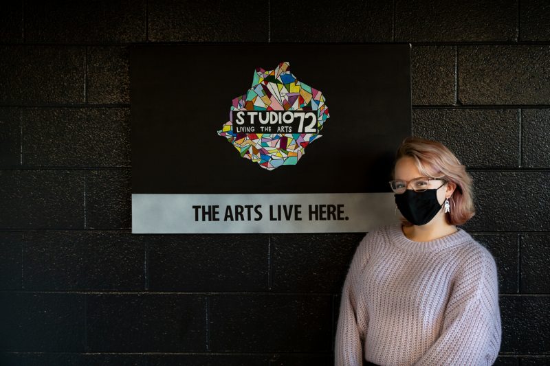 Caillyn Jeffery stands next to a depiction of the Studio 72 Living-Learning Community logo and their tagline: "The arts live here."