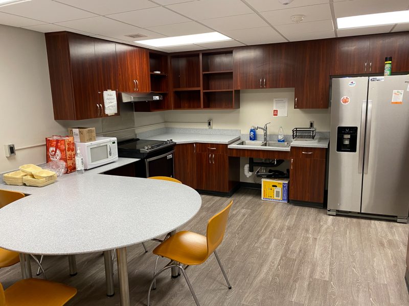 A view of a community kitchen in Hillcrest that includes a fridge, microwave, sink, cabinets, and a small table..
